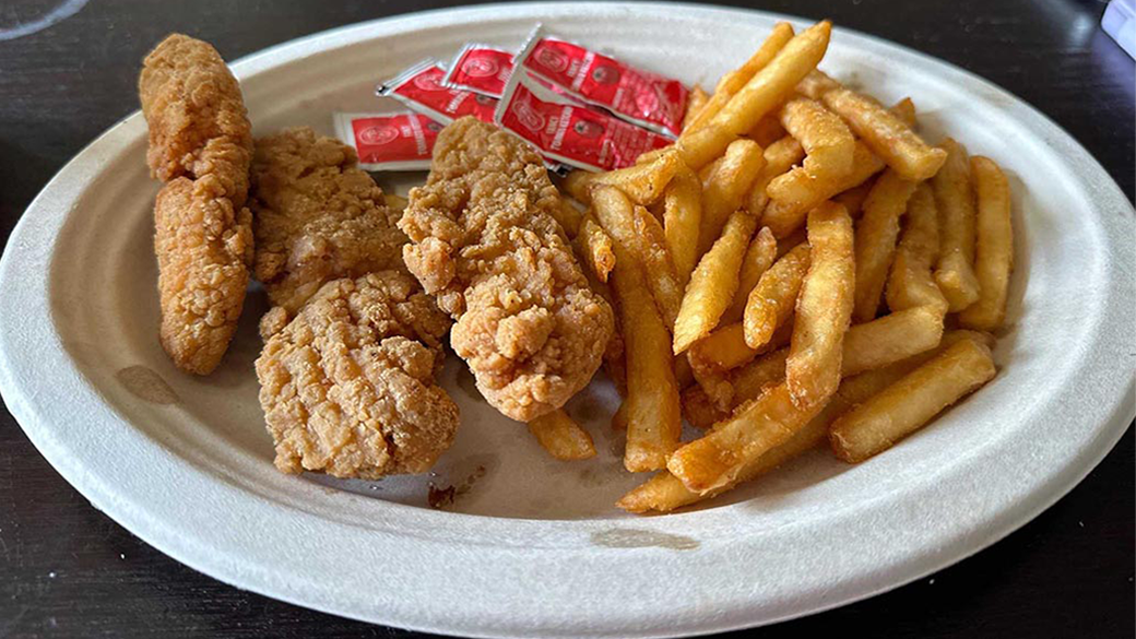 A white paper plate holding crispy, golden-brown chicken strips and a serving of seasoned French fries. Small packets of ketchup are placed beside the chicken strips.