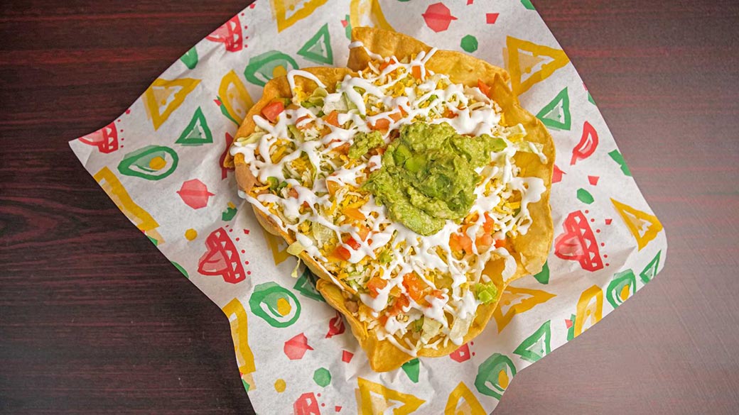 A crispy tortilla bowl filled with shredded lettuce, diced tomatoes, sour cream, shredded cheese, guacamole, and a dollop of salsa.